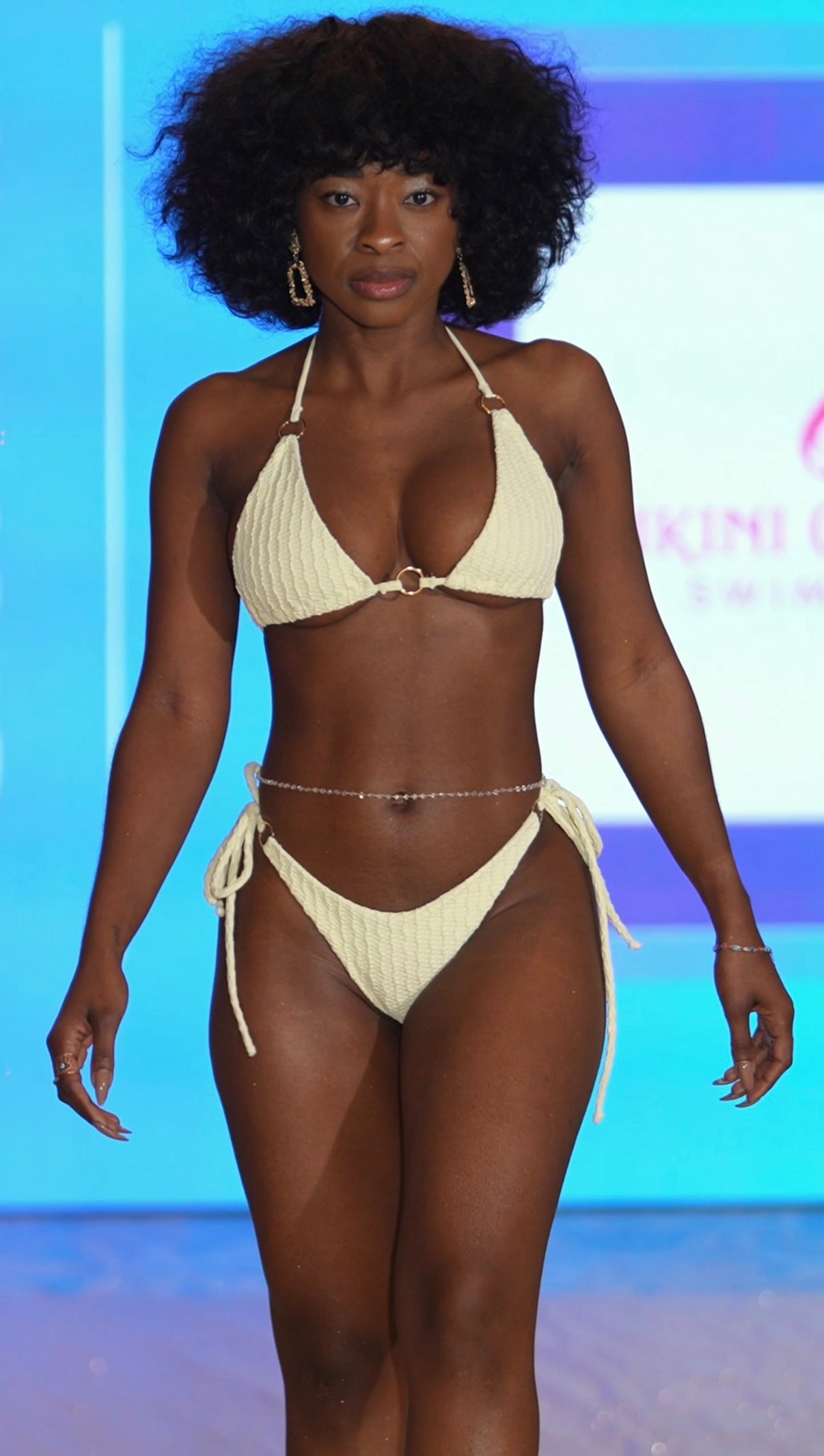 Model wearing a beige bikini on a runway with a blue background